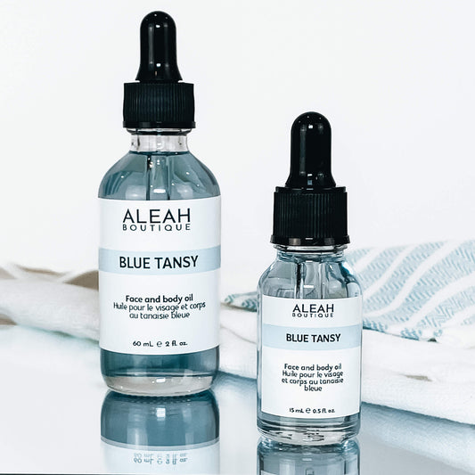 The photo showcases two clear glass dropper bottles of different sizes, both with black caps and labels reading "ALEAH BOUTIQUE BLUE TANSY." They contain a blue-tinted face and body oil, placed on a white surface with a soft blue textile underneath, contributing to a clean and soothing aesthetic.