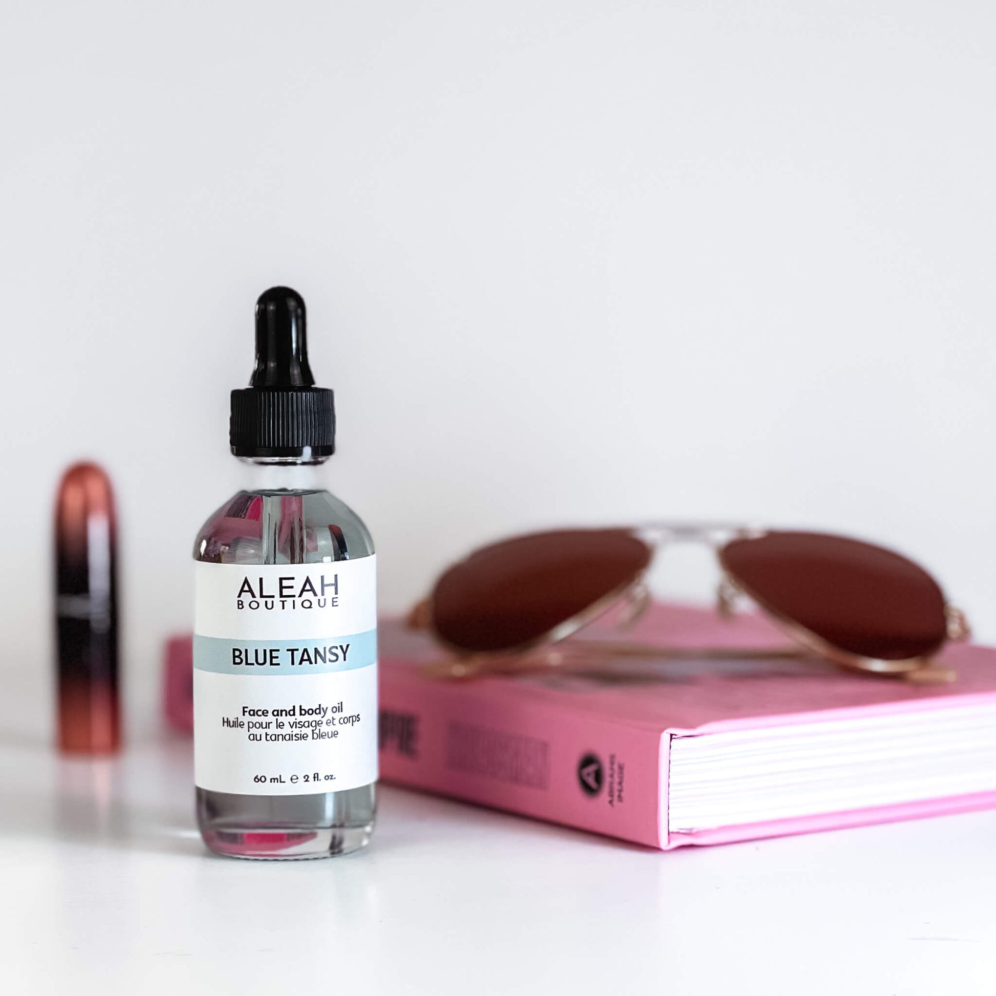 This image features a clear glass dropper bottle with a black cap labeled "ALEAH BOUTIQUE BLUE TANSY." The bottle contains a blue-tinted liquid and is positioned in the foreground against a minimalistic background. Beside the bottle is a pink book and a pair of oversized sunglasses with brown lenses, creating a relaxed, chic ambiance.