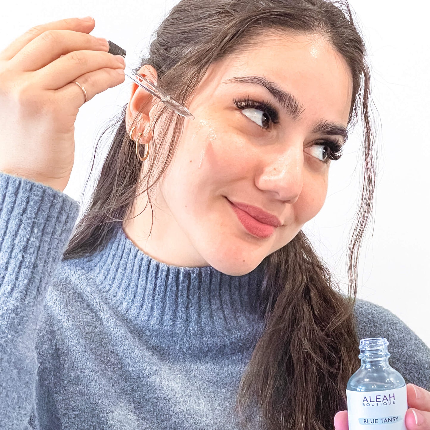 In this image, a person is holding a small glass dropper bottle of "ALEAH BOUTIQUE BLUE TANSY" oil. The individual appears to be applying the blue-tinted oil to their cheek, showcasing the product in use. The person has dark hair, is wearing a blue sweater, and has a happy, content expression, emphasizing the enjoyment of using the product.